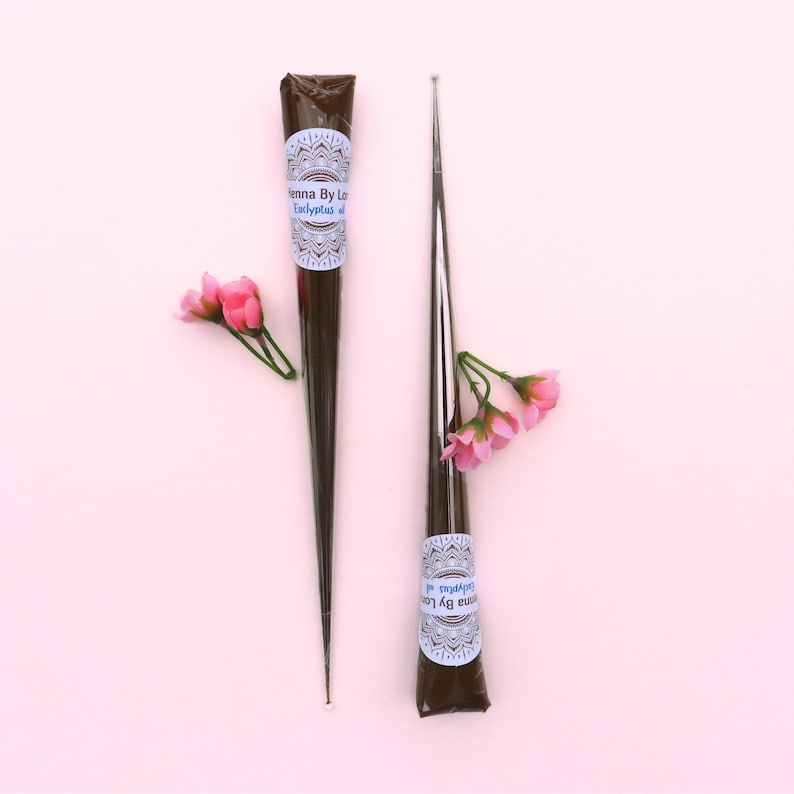 May include: Two henna cones with a floral arrangement on a pink background. The henna cones are dark brown with a white and blue label that reads "Henna By Lo Eucalyptus oil". The cones are next to a metal applicator.