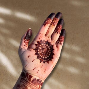 May include: Close-up of a hand adorned with intricate henna designs. The palm features a central circular pattern surrounded by a detailed floral motif. The fingertips are stained a deep brown, and the wrist displays additional henna art.
