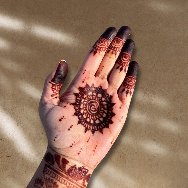 May include: Close-up of a hand adorned with intricate henna designs. The palm features a central circular pattern surrounded by a detailed floral motif. The fingertips are stained a deep brown, and the wrist displays additional henna art.