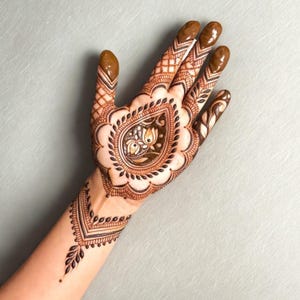 May include: Detailed henna design on a hand, featuring intricate patterns in brown and black. The design includes a central teardrop-shaped motif with floral elements, surrounded by a scalloped border. The fingertips are also adorned with henna.
