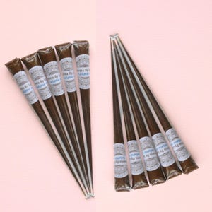 May include: Several henna cones are arranged on a pink surface. The cones are dark brown with white and blue labels that read "Henna By Eucalyptus." The henna cones are packaged in a cone shape.
