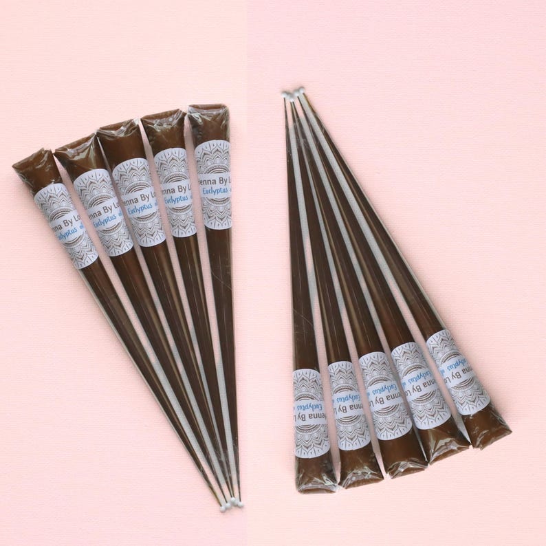 May include: Several henna cones are arranged on a pink surface. The cones are dark brown with white and blue labels that read "Henna By Eucalyptus." The henna cones are packaged in a cone shape.