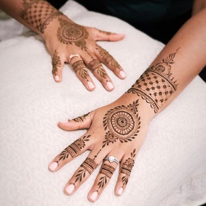 May include: Close-up of hands adorned with intricate henna designs. The henna art features circular and floral patterns, with detailed linework on the fingers and wrists. The skin tone is a warm brown, and the nails are painted with a light pink polish.