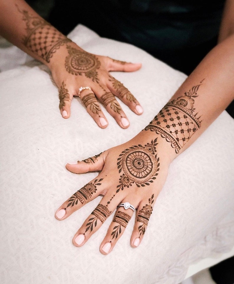 May include: Close-up of hands adorned with intricate henna designs. The henna art features circular and floral patterns, with detailed linework on the fingers and wrists. The skin tone is a warm brown, and the nails are painted with a light pink polish.