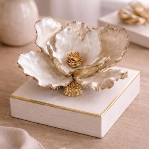 May include: A decorative white and gold floral sculpture. The flower has large, textured petals with gold edges and a gold center. It sits atop a white square base with gold trim. The sculpture is a unique home decor item.