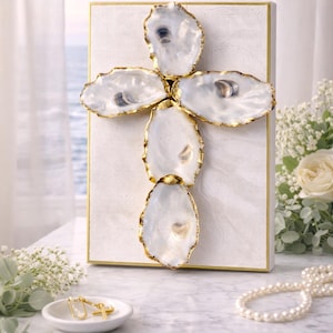 May include: A white canvas cross adorned with five oyster shells with gold-colored edges. The cross is framed in gold and sits on a marble surface. A small dish with gold earrings and a pearl necklace are nearby.