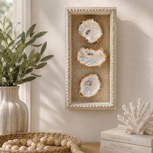 May include: A framed wall art piece featuring three oyster shells with gold-painted edges. The shells are set against a neutral linen background within a rectangular, beaded-edge frame. The artwork evokes a coastal theme.