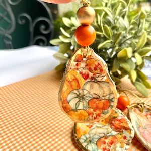 May include: Two gold-leafed oyster shell ornaments with a fall-themed pumpkin and berry design. The ornaments are strung on a gold cord with orange and gold beads.