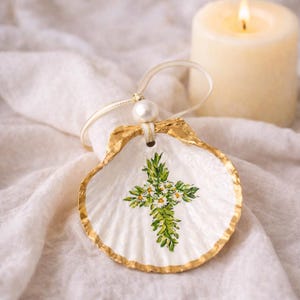 May include: A white seashell ornament with a gold-painted edge and a painted cross design. The cross is decorated with green leaves and small white flowers. A pearl bead and ribbon are attached for hanging.