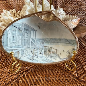 May include: A hand-painted seashell art piece depicting a beach scene with a small house, fence, and birds. The shell is displayed on a gold-colored metal stand. The artwork features shades of blue, white, and brown.