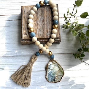 May include: A decorative wooden bead garland with a tassel and an oyster shell pendant. The garland features natural, blue, and gold beads. The pendant has a heron illustration. The garland is displayed on a wooden block.