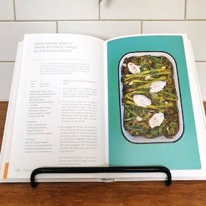 May include: Open cookbook with a recipe for Green Machine Roasted Greens with Basil, Handful Bulgur Wheat & Ricotta. The recipe is shown on the left page and a photo of the finished dish is on the right page. The photo shows a casserole dish filled with green vegetables, ricotta cheese, and herbs.
