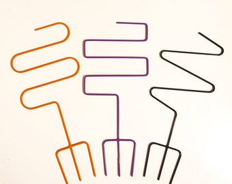 May include: Three metal plant stakes with different designs. The stake on the left is orange and has a wavy design. The stake in the middle is purple and has a straight design. The stake on the right is black and has a zig-zag design. All three stakes have a three-pronged base.