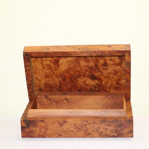 Peut inclure: Une boîte rectangulaire en bois avec un couvercle à charnière, présentant un motif de grain de bois de ronce naturel dans des tons de brun moyen à foncé. L'intérieur de la boîte est visible, montrant une finition en bois brun plus clair.