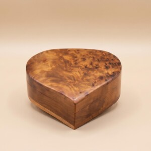 Peut inclure: Une boîte en bois en forme de cœur avec une finition marron chaud. Le couvercle présente un motif de loupe de bois unique. La boîte est probablement utilisée pour ranger des bijoux ou de petits souvenirs.