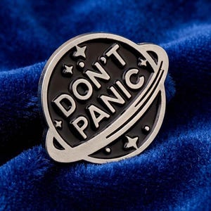 May include: A silver and black enamel pin in the shape of a planet with the words "DON'T PANIC" in white lettering. The pin features stars and a ring around the planet. The pin is set against a blue background.