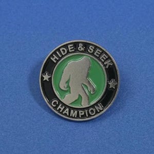 May include: A round, silver-colored pin with a black border and the words "HIDE & SEEK CHAMPION". The pin features a green circle with a silver silhouette of Bigfoot. Two small stars flank the text.