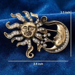 May include: A gold-toned brooch featuring a sun and moon design. The sun has a smiling face and radiating rays, and the moon has a face. Both are adorned with clear rhinestones. The brooch measures 2.4 inches wide and 1.5 inches high.