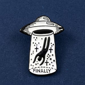 May include: A black and white enamel pin featuring a UFO design. The pin depicts a person being abducted by the UFO, with the word "FINALLY" at the bottom. The pin has a black outline and is set against a dark blue background.