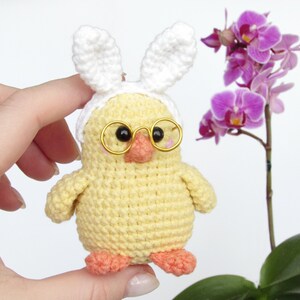 May include: A crocheted yellow chick wearing white bunny ears and gold-rimmed glasses. The chick is holding a small pink flower in its beak.