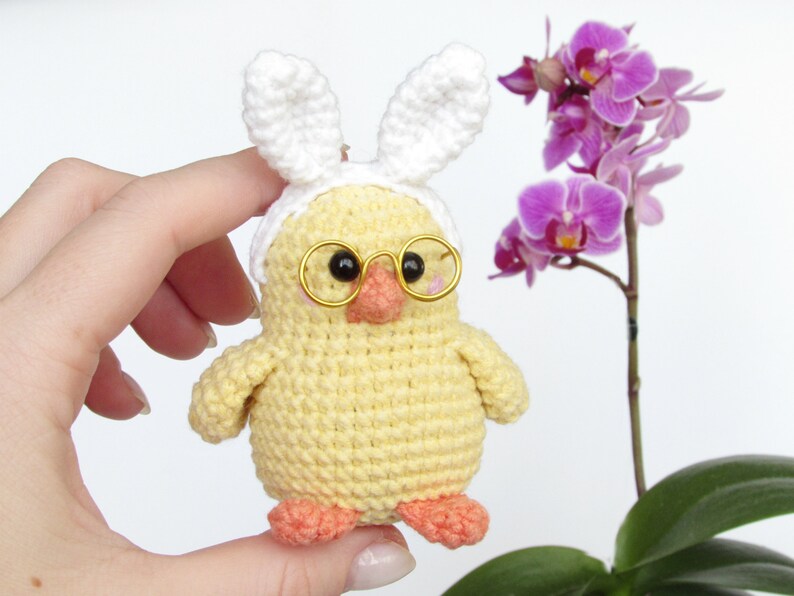 May include: A crocheted yellow chick wearing white bunny ears and gold-rimmed glasses. The chick is holding a small pink flower in its beak.