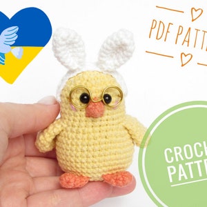 May include: Crochet pattern for a yellow chick wearing bunny ears and glasses. The pattern includes instructions for making the chick, ears, and glasses. The pattern is available as a PDF download. The image also includes a heart with the Ukrainian flag colors and a dove.