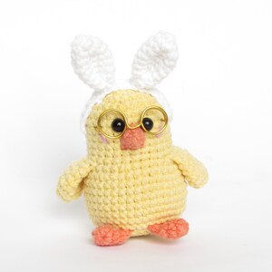 May include: A yellow crocheted chick wearing white bunny ears and gold-rimmed glasses. The chick has a pink beak and orange feet.