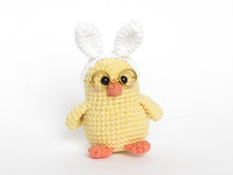 May include: A yellow crocheted chick wearing white bunny ears and gold-rimmed glasses. The chick has a pink beak and orange feet.