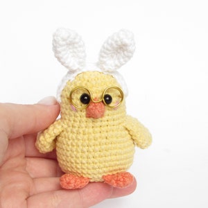May include: A small yellow crocheted chick wearing white bunny ears and gold-rimmed glasses. The chick has a small orange beak and pink feet.