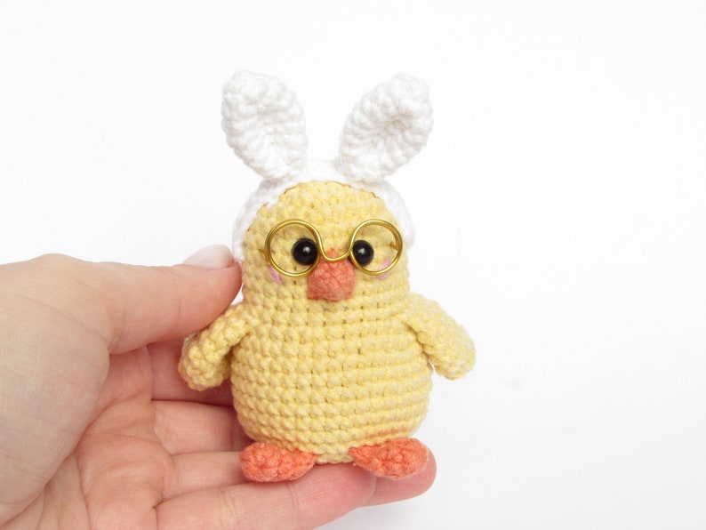 May include: A small yellow crocheted chick wearing white bunny ears and gold-rimmed glasses. The chick has a small orange beak and pink feet.