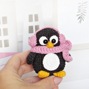 May include: A small crocheted penguin wearing a pink scarf and earmuffs. The penguin has black and white fur and yellow feet.
