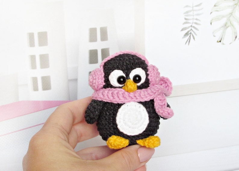May include: A small crocheted penguin wearing a pink scarf and earmuffs. The penguin has black and white fur and yellow feet.
