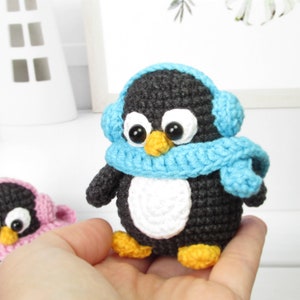 May include: Two small crocheted penguin toys wearing blue and pink scarves and earmuffs. The penguins have black bodies, white bellies, and orange beaks.