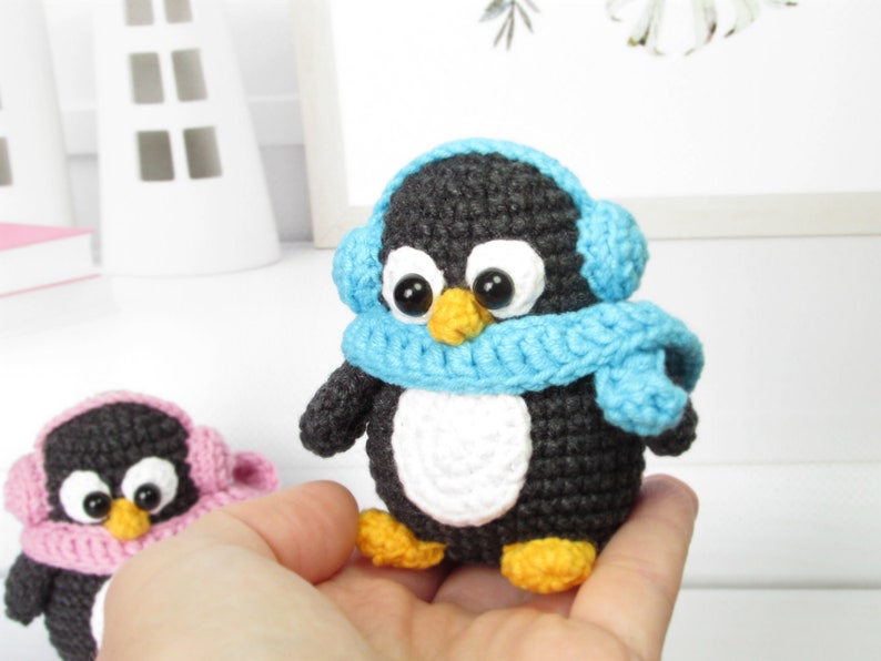 May include: Two small crocheted penguin toys wearing blue and pink scarves and earmuffs. The penguins have black bodies, white bellies, and orange beaks.