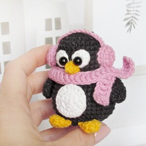 May include: A small crocheted penguin wearing a pink scarf and earmuffs. The penguin is black and white with a white belly and black eyes.