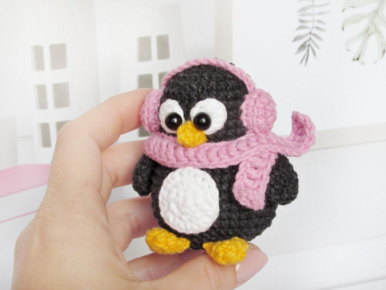 May include: A small crocheted penguin wearing a pink scarf and earmuffs. The penguin is black and white with a white belly and black eyes.