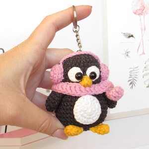 May include: A crocheted penguin keychain wearing a pink scarf and earmuffs. The penguin has black and white fur and a white belly. The keychain is attached to a silver chain.