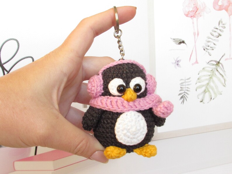 May include: A crocheted penguin keychain wearing a pink scarf and earmuffs. The penguin has black and white fur and a white belly. The keychain is attached to a silver chain.