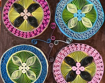 May include: Four circular paper quilling art pieces with a floral design. Each piece has a different color scheme: pink, blue, green, and pink. The pieces are arranged in a square pattern on a brown background.