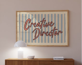 May include: A wooden dresser with three drawers and two doors. The dresser is brown and has a light wood finish. A white lamp and a bowl are on top of the dresser. A framed print with the text "Creative Director" is hanging on the wall above the dresser.