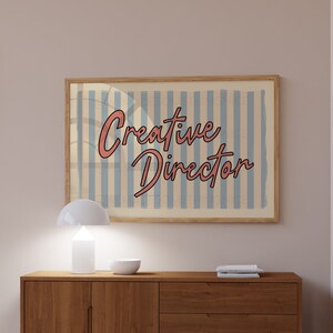 May include: A wooden dresser with three drawers and two doors. The dresser is brown and has a light wood finish. A white lamp and a bowl are on top of the dresser. A framed print with the text "Creative Director" is hanging on the wall above the dresser.