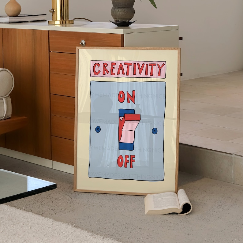 May include: A framed art print with a light beige background. The artwork features a blue light switch with the words "ON" and "OFF" in red. Above the switch, the word "CREATIVITY" is written in red. The print is in a light brown wooden frame.