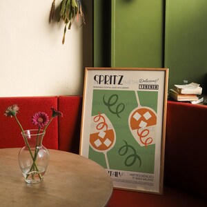 Pode incluir: Um p&ocirc;ster emoldurado com fundo verde e ilustra&ccedil;&otilde;es laranja e brancas de coquet&eacute;is. O p&ocirc;ster tem o texto "SPRITZ Delicious! 1998" e "@Wedesomethingnice" em texto preto. O p&ocirc;ster est&aacute; pendurado em uma parede verde ao lado de um banco vermelho.