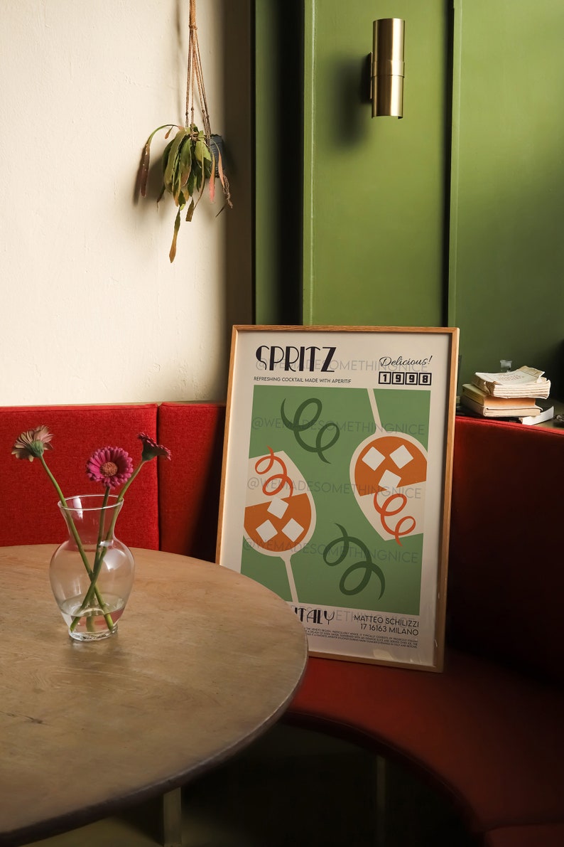 Pode incluir: Um p&ocirc;ster emoldurado com fundo verde e ilustra&ccedil;&otilde;es laranja e brancas de coquet&eacute;is. O p&ocirc;ster tem o texto "SPRITZ Delicious! 1998" e "@Wedesomethingnice" em texto preto. O p&ocirc;ster est&aacute; pendurado em uma parede verde ao lado de um banco vermelho.