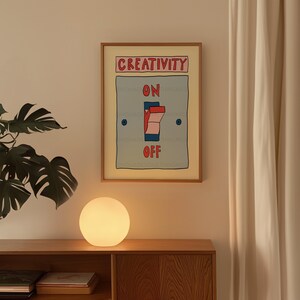 May include: A framed artwork with the word "CREATIVITY" above a light switch illustration that reads "ON" and "OFF". A round, glowing lamp sits on a wooden cabinet below the artwork. A large green plant and a white curtain are also visible.