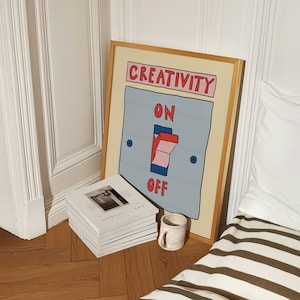 May include: A framed art print with a light beige border features a light blue switch with the words "CREATIVITY" above and "ON" and "OFF" below. A stack of white books and a small ceramic mug sit on the wooden floor.