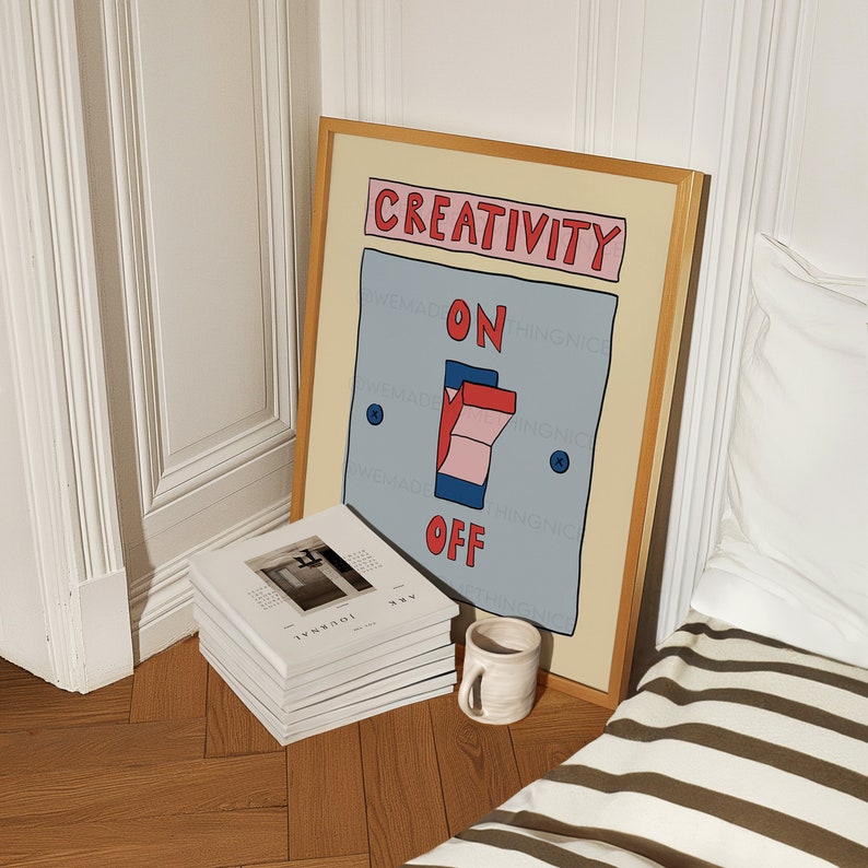May include: A framed art print with a light beige border features a light blue switch with the words "CREATIVITY" above and "ON" and "OFF" below. A stack of white books and a small ceramic mug sit on the wooden floor.