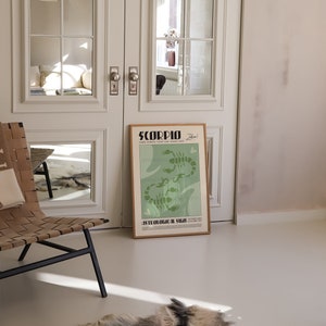 Op de afbeelding: Een groene en witte poster met het woord "SCORPIO" in vetgedrukte zwarte letters. De poster toont een gestileerde illustratie van twee schorpioenen met de tekst "Astrologisch teken" eronder. De poster is ingelijst in een licht houten lijst en leunt tegen een witte deur.