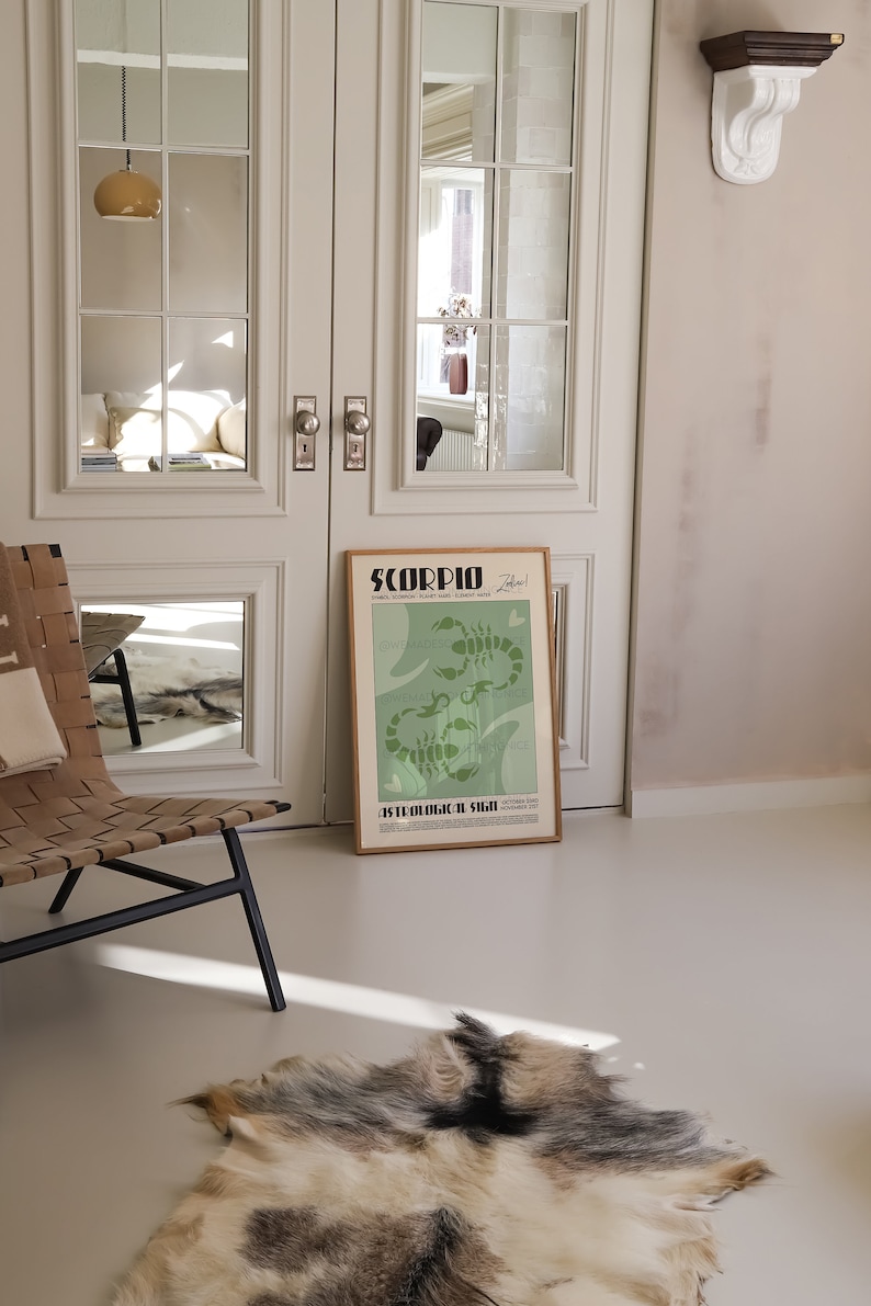 Op de afbeelding: Een groene en witte poster met het woord "SCORPIO" in vetgedrukte zwarte letters. De poster toont een gestileerde illustratie van twee schorpioenen met de tekst "Astrologisch teken" eronder. De poster is ingelijst in een licht houten lijst en leunt tegen een witte deur.