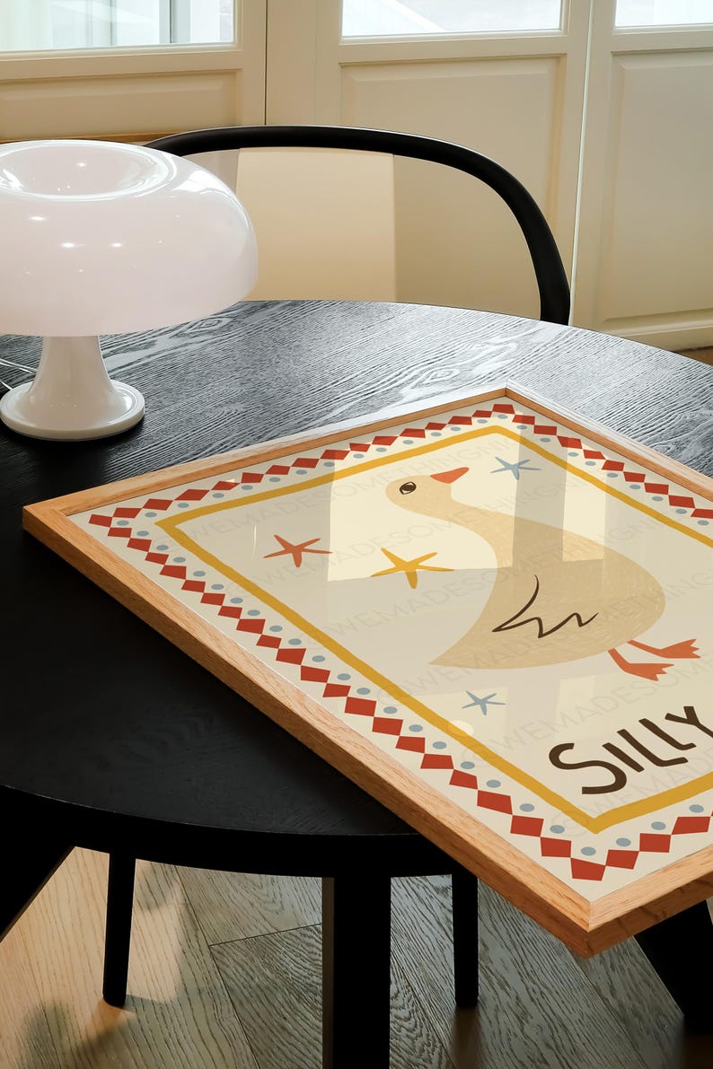 Puede incluir: Una impresi&oacute;n enmarcada con un fondo amarillo que presenta un ganso de dibujos animados con la palabra "Silly" escrita debajo. El marco es de madera y tiene un borde con un patr&oacute;n rojo, blanco y azul.